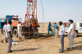 Nine people on sandy ground with a truck and drilling equipment